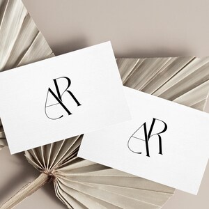 May include: Two white business cards with a black monogram logo of the letters "AR" in a geometric design. The cards are placed on top of two dried palm leaves.