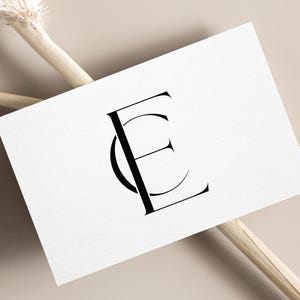May include: A white business card with the black intertwined letters "CE" is displayed at an angle. The card rests on a beige surface, with dried floral stems crossing behind it. The overall aesthetic is minimalist and elegant.
