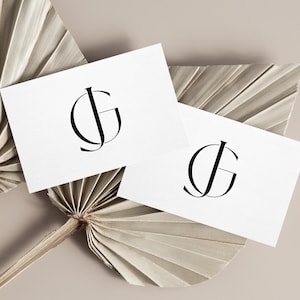 May include: Two white business cards with a black "JG" monogram are displayed on a beige background with dried palm leaves. The cards are slightly angled, showcasing the elegant design.