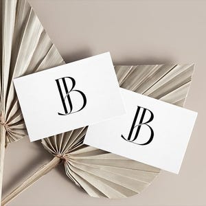 May include: Two white cards with a black stylized letter "B" logo on each. The cards are on a beige background with two dried palm leaves.