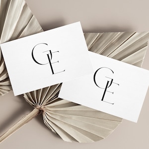 Puede incluir: Dos tarjetas de visita blancas con las letras "GE" en negro, con una fuente elegante. Están dispuestas sobre un fondo beige con hojas de palma secas. El diseño es minimalista y sofisticado, centrado en la tipografía.