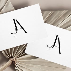 Pode incluir: Dois cartões brancos com as letras "JA" em preto, estilizadas com o "J" sobreposto ao "A". Os cartões estão sobre um fundo castanho claro.