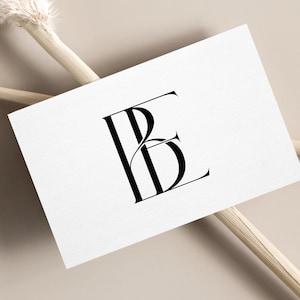 May include: A white business card with a black monogram logo featuring the letters "BE" in a stylized, elegant font. The card rests on a neutral beige surface, accented by dried floral stems.