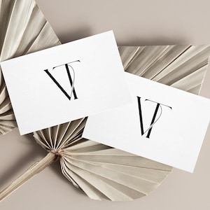 May include: Two white business cards with the intertwined letters "VT" in black, resting on a dried palm leaf and a neutral-toned background. The minimalist design and neutral colors suggest a sophisticated brand identity.