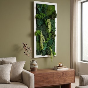 May include: A rectangular wall art piece with a white frame, filled with various shades of green moss and plants. The artwork hangs on a green wall above a wooden cabinet. A vase with decorative branches and a sofa are in the foreground.