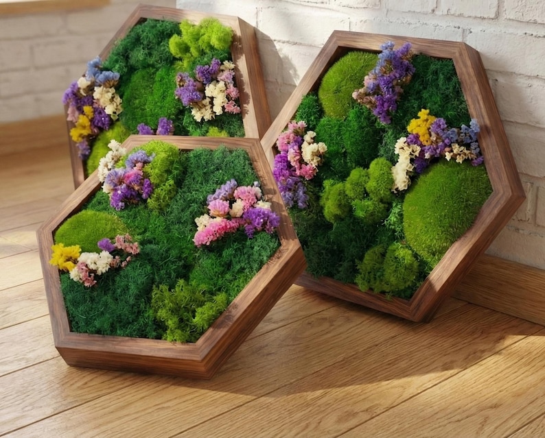 Three handcrafted hexagonal botanical wall art pieces in brown alder wood frames, resting on a wooden floor. Each panel features vibrant preserved green moss and clusters of purple, pink, and yellow flowers.