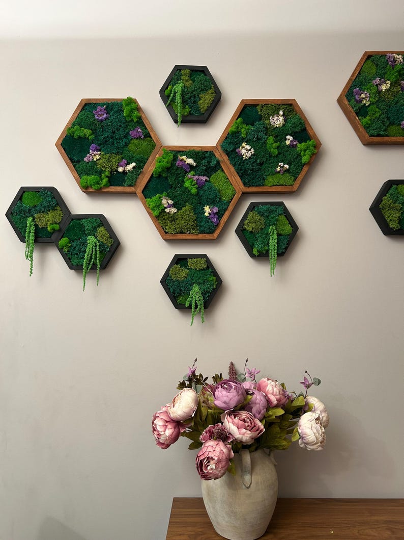 Wall arrangement of multiple hexagonal wooden frames filled with preserved green moss, colorful flowers, and hanging amaranth, displayed above a vase of pink peonies.