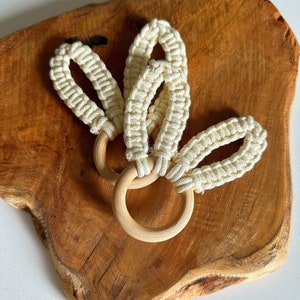 Könnte beinhalten: Drei weiße gehäkelte Schlaufen, die an zwei Holzringen befestigt sind. Die Ringe sind aus Naturholz und haben eine glatte Oberfläche. Die gehäkelten Schlaufen bestehen aus einer weichen, natürlichen Faser.