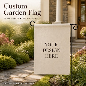 May include: A rectangular, double-sided, beige garden flag featuring the dark brown text "YOUR DESIGN HERE" centered on the fabric. The flag is suspended from a black metal garden flag stand with decorative scrollwork at the top corners.