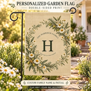 May include: A rectangular beige garden flag featuring a double-sided print of a floral wreath made of green leaves, olives, and white daisies. The centre displays the letter H, the text THE HARRISON FAMILY, and WELCOME TO OUR GARDEN.