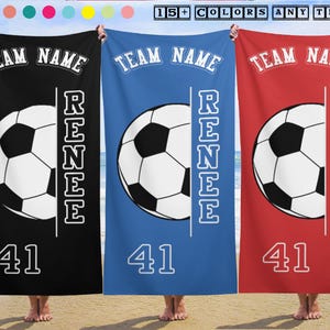 May include: Three personalized soccer towels in black, blue, and red. Each towel features a soccer ball graphic, the text "TEAM NAME", a name, and the number "41". The towels are displayed on a beach.