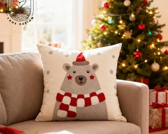 Può includere: Un cuscino decorativo color crema con un orso grigio che indossa una sciarpa a scacchi rossa e bianca e un cappello rosso. L'orso ha accenti di stella rossa ed è centrato sul cuscino. Il cuscino è su un divano beige davanti a un albero di Natale.