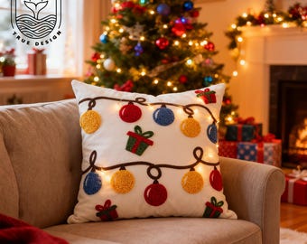 Può includere: Un cuscino bianco decorato con ornamenti natalizi ricamati e scatole regalo. Il cuscino presenta ornamenti a forma di lampadina rossi, blu e gialli e scatole regalo verdi. Il cuscino è su un divano beige davanti a un albero di Natale decorato.