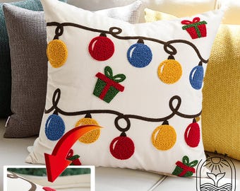 Può includere: Cuscino quadrato bianco con ricami di luci natalizie e confezioni regalo rosse, gialle e blu. Il cuscino è su un divano bianco con altri cuscini sullo sfondo.