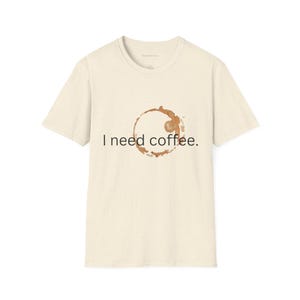 Peut inclure: Un t-shirt beige avec une tache de café et le texte "J'ai besoin de café."