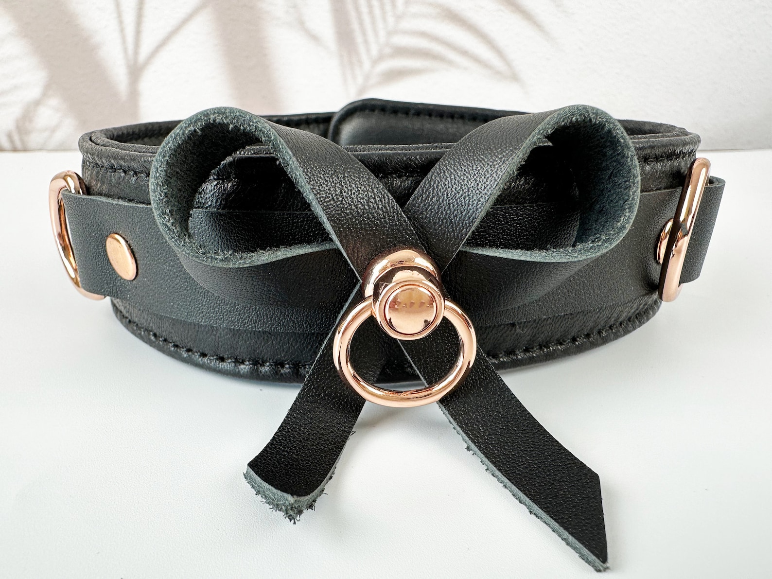 Black leather collar with rose gold hardware and bow detail laid flat on white surface, stylish and elegant bdsm accessory.