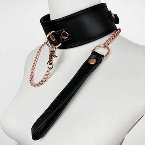 Könnte beinhalten: Ein schwarzes Lederhalsband mit einer roségoldenen Kette und einer passenden Leine. Das Halsband hat roségoldene Akzente und die Leine einen roségoldenen Ring.