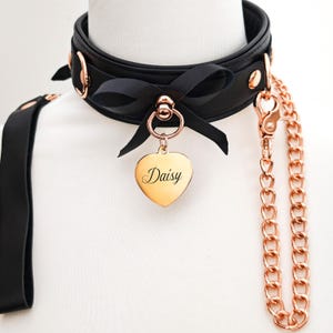 Peut inclure: Collier en cuir noir avec des éléments en or rose, un nœud noir et une étiquette en forme de cœur gravée "Daisy". Comprend une laisse à chaîne en or rose assortie.