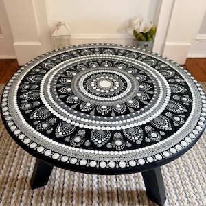 May include: A round black wooden table featuring a detailed hand-painted mandala design in shades of white, gray, and silver. The tabletop displays concentric circles of dots forming intricate patterns, supported by three sturdy black legs.