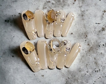 May include: A set of ten press-on nails with a translucent nude base, featuring gold-toned metallic chains, clear rhinestones, and large amber-colored faceted gems arranged in decorative patterns across the surface of each individual nail.