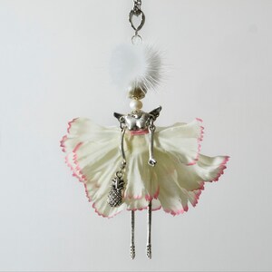 May include: A silver charm necklace featuring a delicate figurine of a woman wearing a white fur hat and a white dress with pink edges. The woman is holding a small silver pineapple charm.