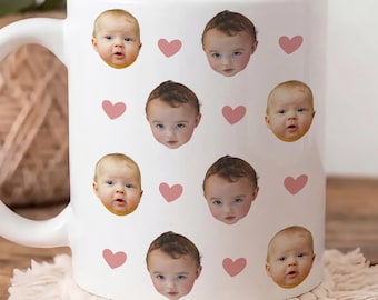 May include: A white ceramic mug featuring a repeating pattern of two different baby faces and small pink hearts. The mug has a curved handle on the left side and a smooth, glossy finish, displaying the printed faces of infants in a grid-like arrangement.