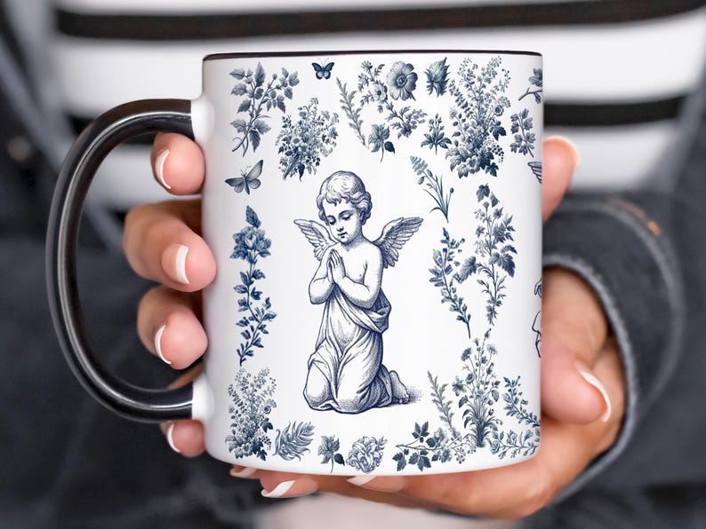 May include: White ceramic mug with a black handle and rim, featuring a blue and white floral design with an angel illustration. The mug is held by a person.