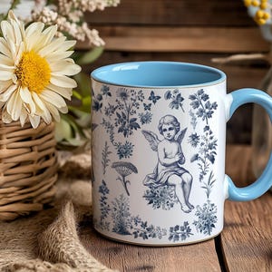 May include: White ceramic mug with a light blue interior and handle. The mug features a blue and white floral design with an angel illustration. The mug is sitting on a wooden surface, with flowers in the background.