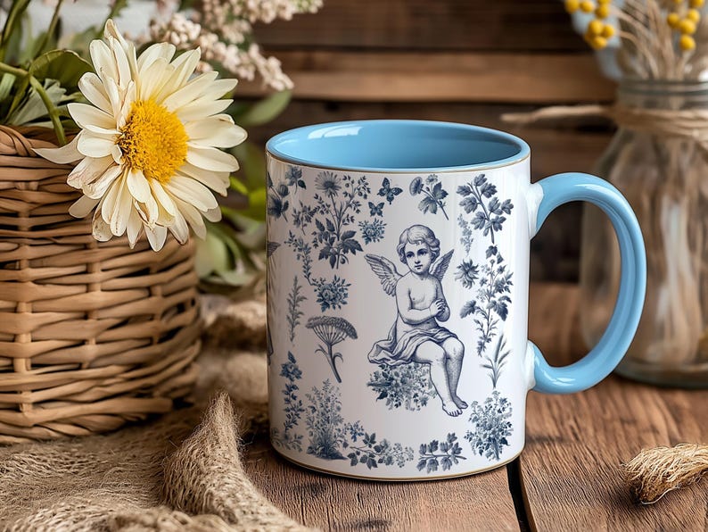 May include: White ceramic mug with a light blue interior and handle. The mug features a blue and white floral design with an angel illustration. The mug is sitting on a wooden surface, with flowers in the background.