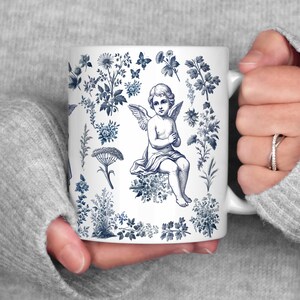 May include: White ceramic mug featuring a blue and white floral design with a central illustration of an angel. The mug has a curved handle and is held by a person wearing a gray sweater. The design includes flowers, butterflies, and botanical elements.