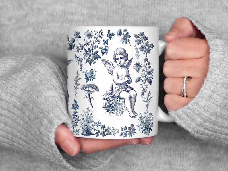 May include: White ceramic mug featuring a blue and white floral design with a central illustration of an angel. The mug has a curved handle and is held by a person wearing a gray sweater. The design includes flowers, butterflies, and botanical elements.