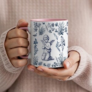 May include: White ceramic mug with a pink interior and handle. The mug features a blue and white illustration of an angel kneeling in prayer, surrounded by floral and butterfly motifs. A decorative item.