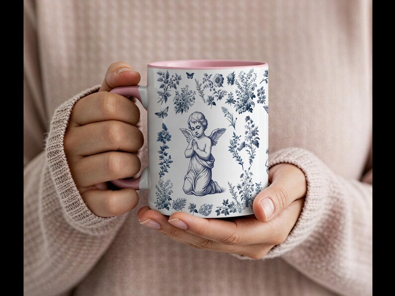 May include: White ceramic mug with a pink interior and handle. The mug features a blue and white illustration of an angel kneeling in prayer, surrounded by floral and butterfly motifs. A decorative item.