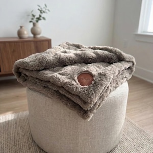 May include: A plush, taupe-colored blanket is draped over a round, upholstered ottoman. The blanket has a soft, textured appearance and features a circular leather patch with the text "mama family". The ottoman sits on a neutral-toned rug.