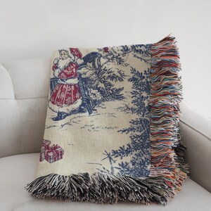 May include: A woven Christmas throw blanket featuring a festive scene with Santa Claus, gifts, and trees in blue and red. The blanket has a cream background and is trimmed with a colorful fringe.