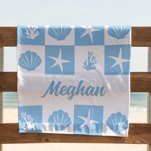 Puede incluir: Una toalla de playa blanca con un patr&oacute;n de cuadros azules que presenta conchas marinas, estrellas de mar y corales. El nombre "Meghan" est&aacute; escrito en letras cursivas azules.