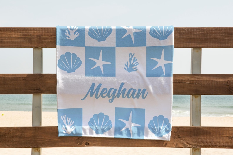Puede incluir: Una toalla de playa blanca con un patr&oacute;n de cuadros azules que presenta conchas marinas, estrellas de mar y corales. El nombre "Meghan" est&aacute; escrito en letras cursivas azules.
