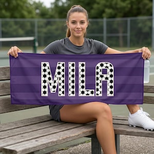 May include: A purple sports towel with the name "MILA" in white letters filled with soccer ball patterns. The towel has horizontal stripes and is held by a person sitting on a wooden bench.