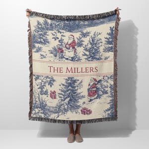May include: A cream-colored throw blanket with a blue and red Christmas toile design. The blanket features a forest scene with Santa Claus figures and gift boxes. The words "THE MILLERS" are printed in red.