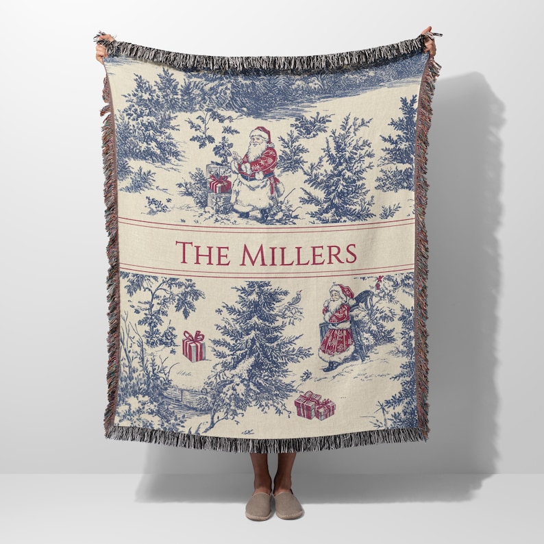 May include: A cream-colored throw blanket with a blue and red Christmas toile design. The blanket features a forest scene with Santa Claus figures and gift boxes. The words "THE MILLERS" are printed in red.