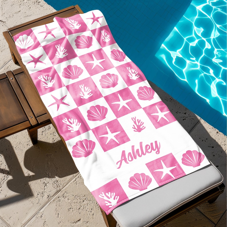 Puede incluir: Toalla de playa rosa y blanca con un patr&oacute;n a cuadros con conchas, estrellas de mar y coral. La toalla tiene el nombre "Ashley" escrito en cursiva.