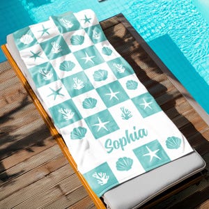 Puede incluir: Toalla de playa blanca con un patr&oacute;n a cuadros en azul turquesa. La toalla presenta dise&ntilde;os de conchas marinas, estrellas de mar y corales. El nombre "Sophia" est&aacute; escrito en una fuente cursiva azul turquesa.