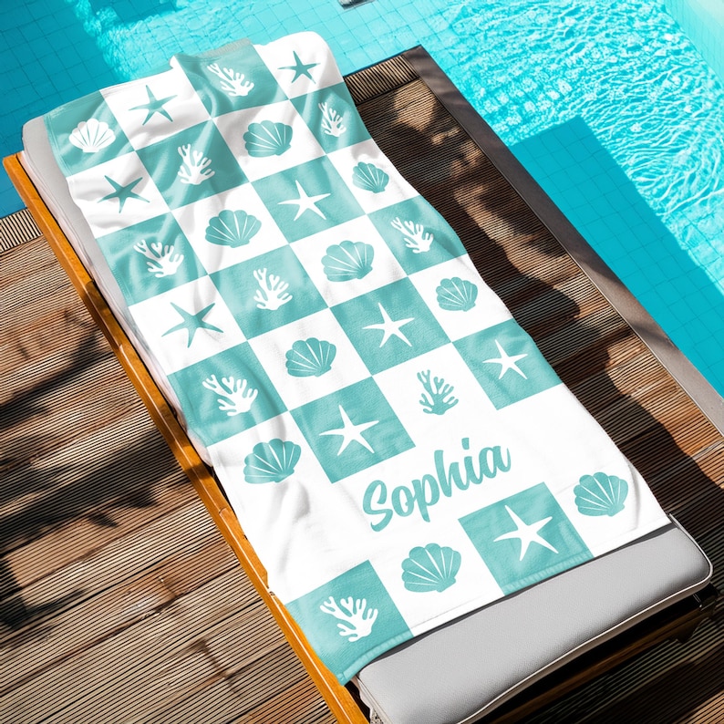 Puede incluir: Toalla de playa blanca con un patr&oacute;n a cuadros en azul turquesa. La toalla presenta dise&ntilde;os de conchas marinas, estrellas de mar y corales. El nombre "Sophia" est&aacute; escrito en una fuente cursiva azul turquesa.