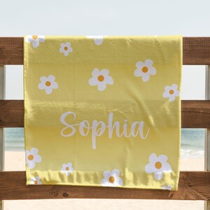 May include: Yellow beach towel with a white daisy pattern and the name "Sophia" written in white cursive. The towel is hanging on a wooden fence.
