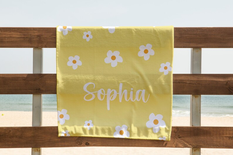 May include: Yellow beach towel with a white daisy pattern and the name "Sophia" written in white cursive. The towel is hanging on a wooden fence.