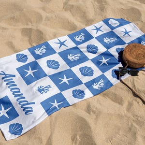 Puede incluir: Una toalla de playa blanca con un patr&oacute;n de cuadros azul y blanco. La toalla presenta dise&ntilde;os de conchas marinas, estrellas de mar y corales. La palabra "Amanda" est&aacute; escrita en cursiva azul en la toalla. Un bolso tejido marr&oacute;n y gafas de sol est&aacute;n encima de la toalla.