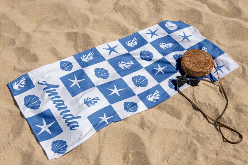 Puede incluir: Una toalla de playa blanca con un patr&oacute;n de cuadros azul y blanco. La toalla presenta dise&ntilde;os de conchas marinas, estrellas de mar y corales. La palabra "Amanda" est&aacute; escrita en cursiva azul en la toalla. Un bolso tejido marr&oacute;n y gafas de sol est&aacute;n encima de la toalla.