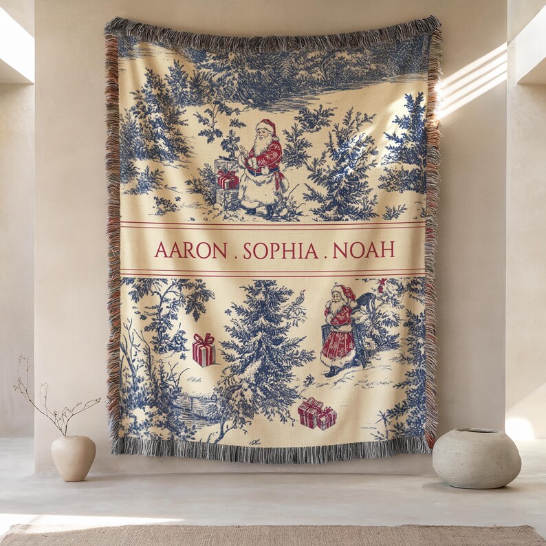 May include: Cream-colored Christmas throw blanket with a blue and red toile design. The blanket features a winter scene with Santa Claus figures, trees, and gift boxes. The names "AARON. SOPHIA. NOAH" are printed in red across the center. The blanket has a fringed edge.