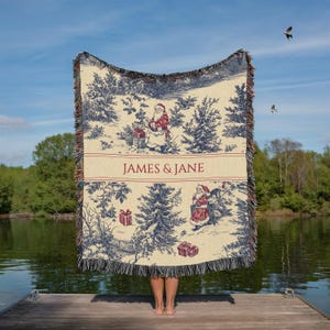 May include: A cream-colored Christmas throw blanket with a blue and red toile design featuring Santa Claus and pine trees. The blanket has a fringed edge and the text "JAMES & JANE" in red.