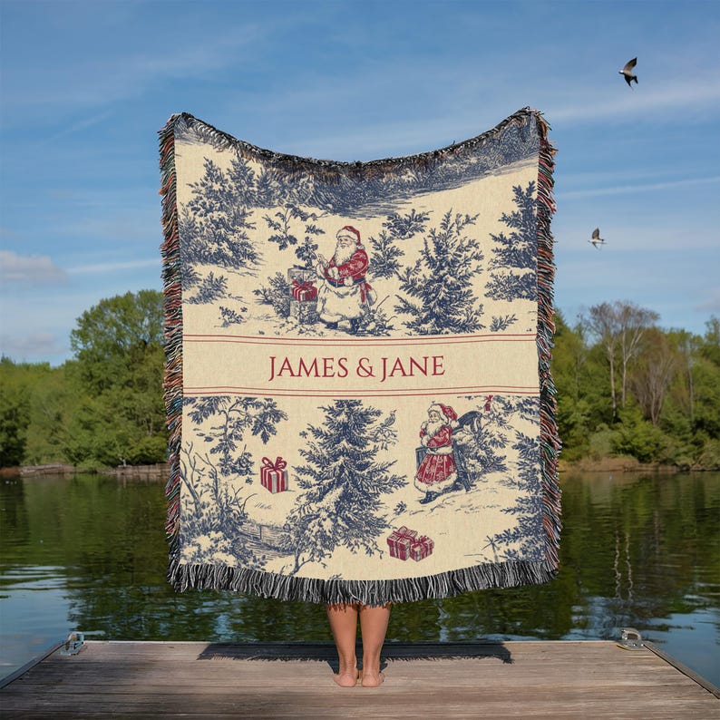 May include: A cream-colored Christmas throw blanket with a blue and red toile design featuring Santa Claus and pine trees. The blanket has a fringed edge and the text "JAMES & JANE" in red.